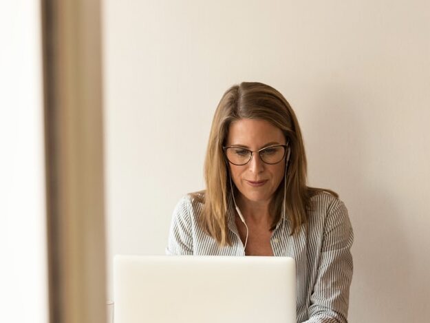 woman wearing grey striped dress shirt sitting down near brown wooden table in front of white laptop computer
