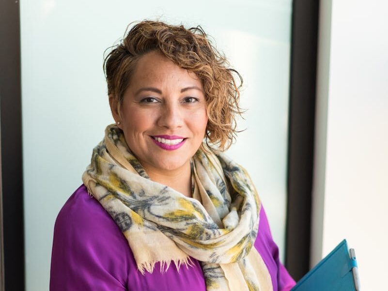 Smiling woman in a purple dress and scarf holding a tablet indoors.