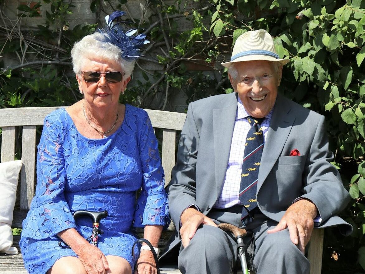 A happy elderly couple sitting together on a garden bench, smiling outdoors.