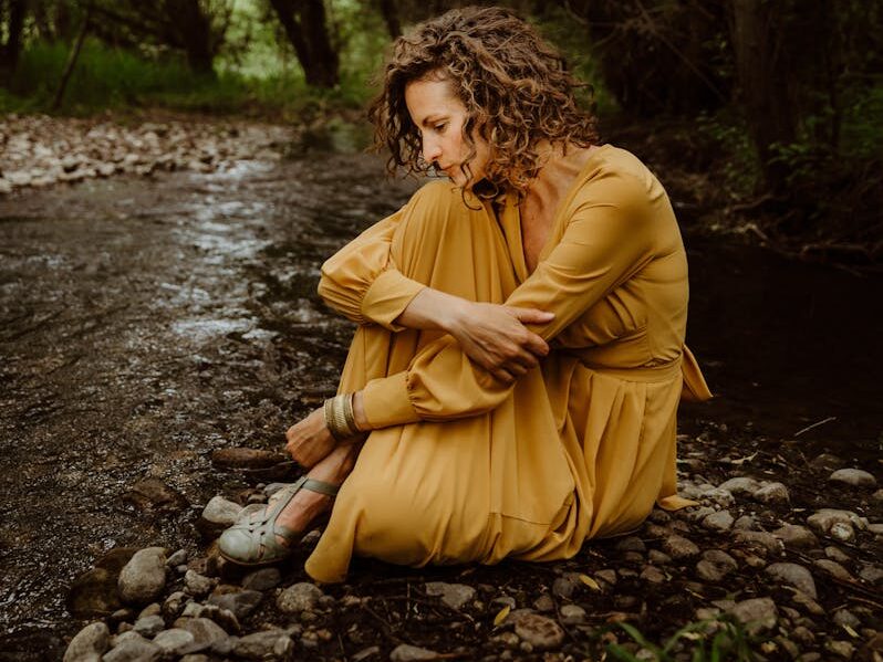A woman in yellow sits pensively by a forest stream, reflecting amidst nature.