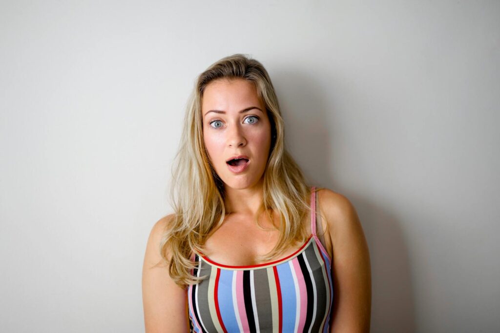 A young woman with a shocked expression stands against a plain white wall.