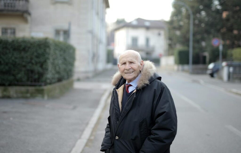 A cheerful elderly man in a coat smiling while standing outdoors on a city street.
