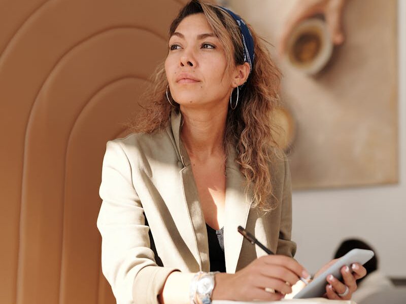 Confident woman in blazer taking notes with pen and smartphone in cafe.