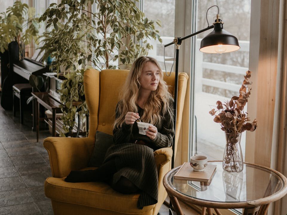 Woman sitting in a cozy armchair by a window, surrounded by houseplants, enjoying a quiet moment indoors.