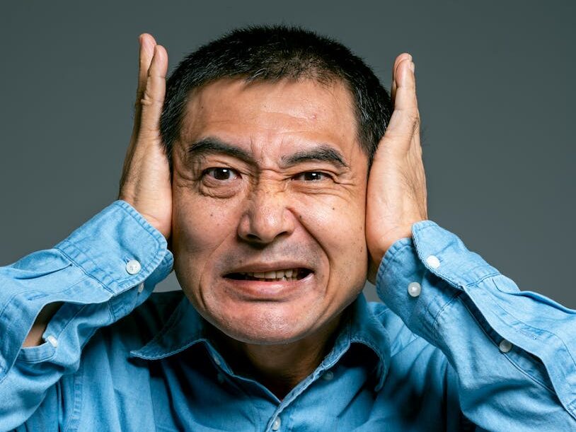 Close-up portrait of an Asian man in a blue shirt expressing frustration by holding his ears.