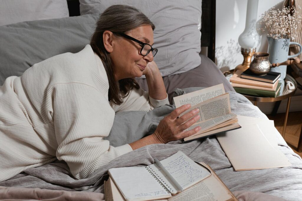 Senior woman reading comfortably in bed with eyeglasses and a cozy blanket, enjoying a serene moment.