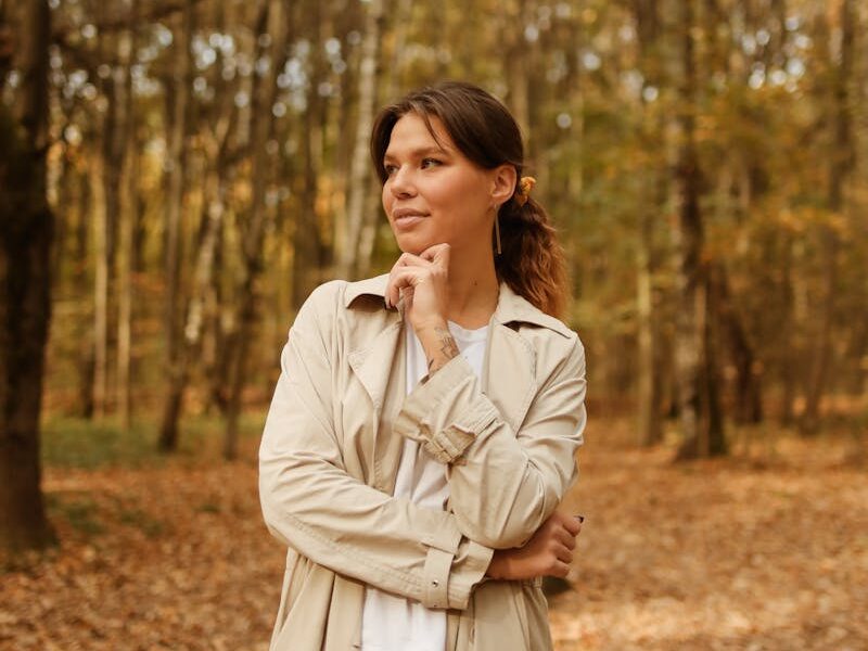 Woman in a trench coat standing thoughtfully in an autumn forest scene.