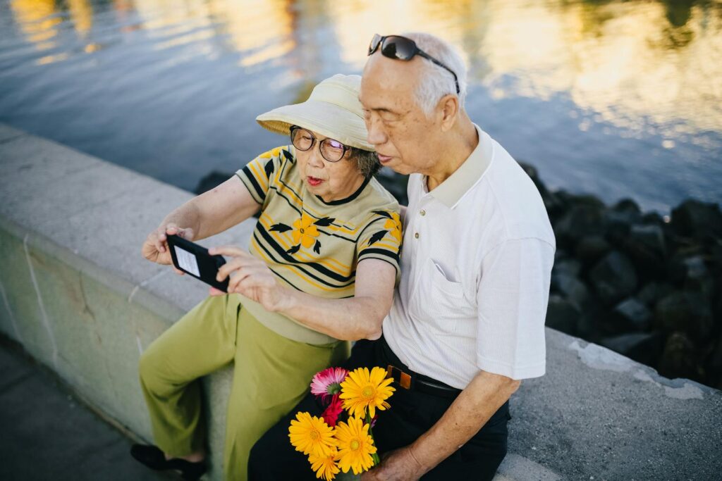 An elderly Asian couple takes a cheerful selfie by a waterfront while enjoying a sunny day.