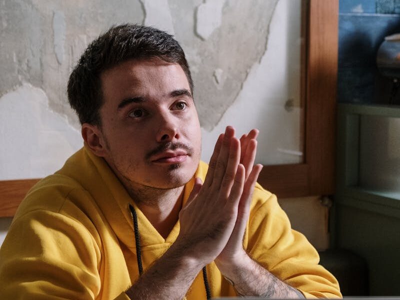 Man in yellow hoodie thinking deeply next to a laptop indoors.