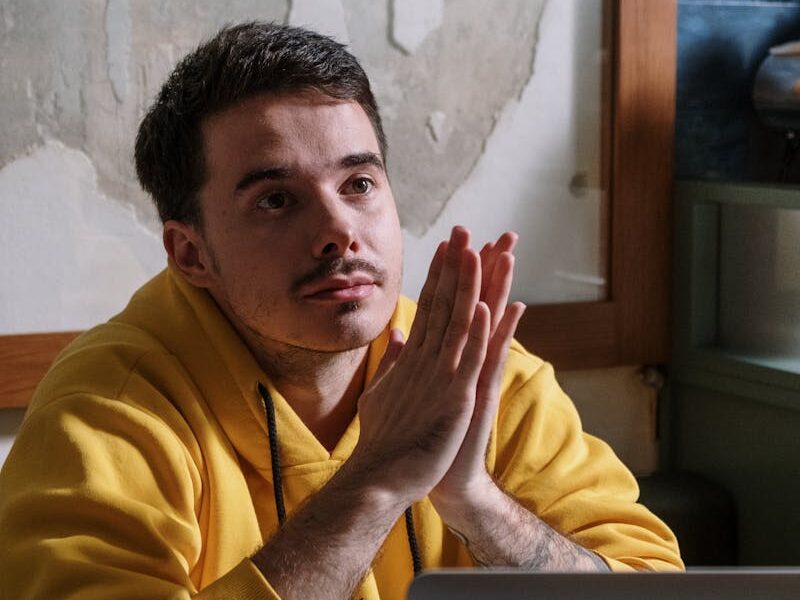 Man in yellow hoodie thinking deeply next to a laptop indoors.