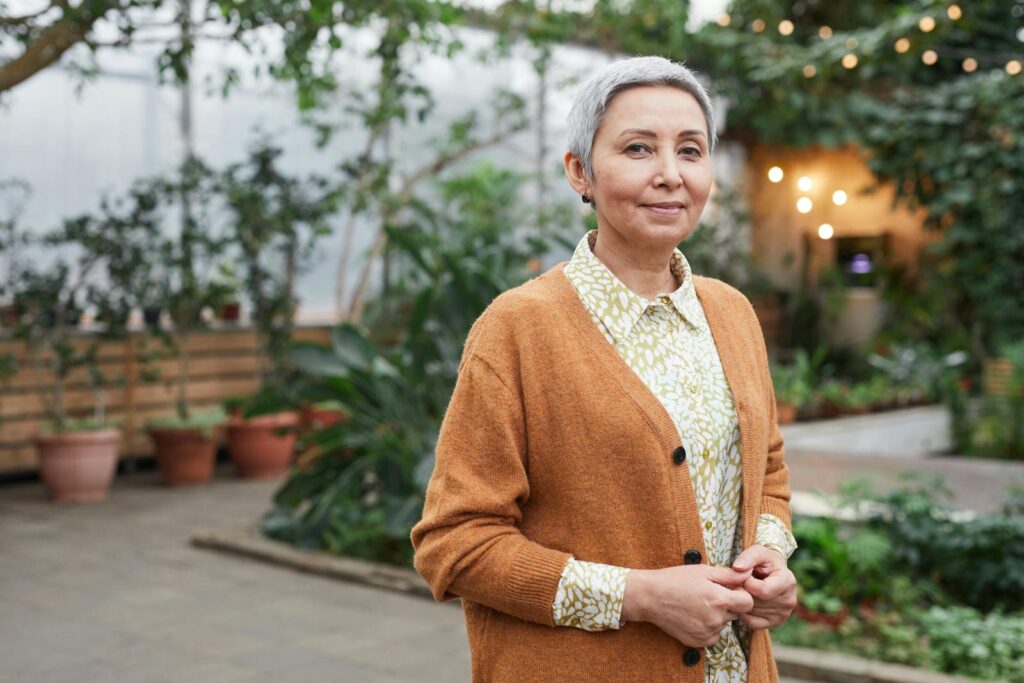 A gracefully smiling senior woman in a stylish cardigan poses in a serene indoor greenhouse.