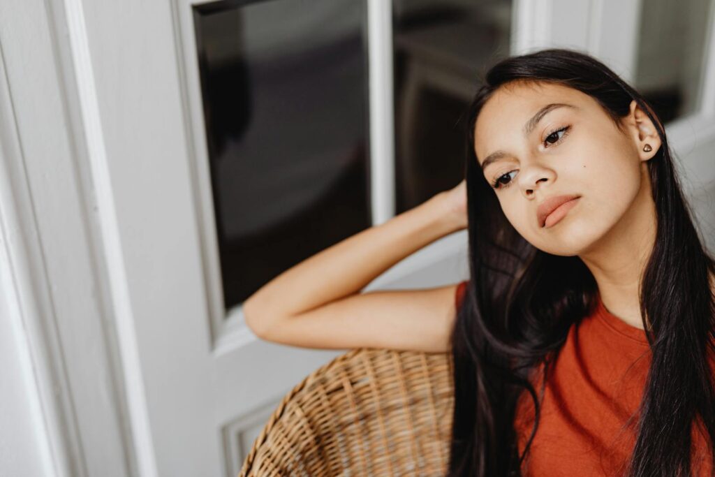 Young woman with long dark hair sitting pensively in a wicker chair indoors.