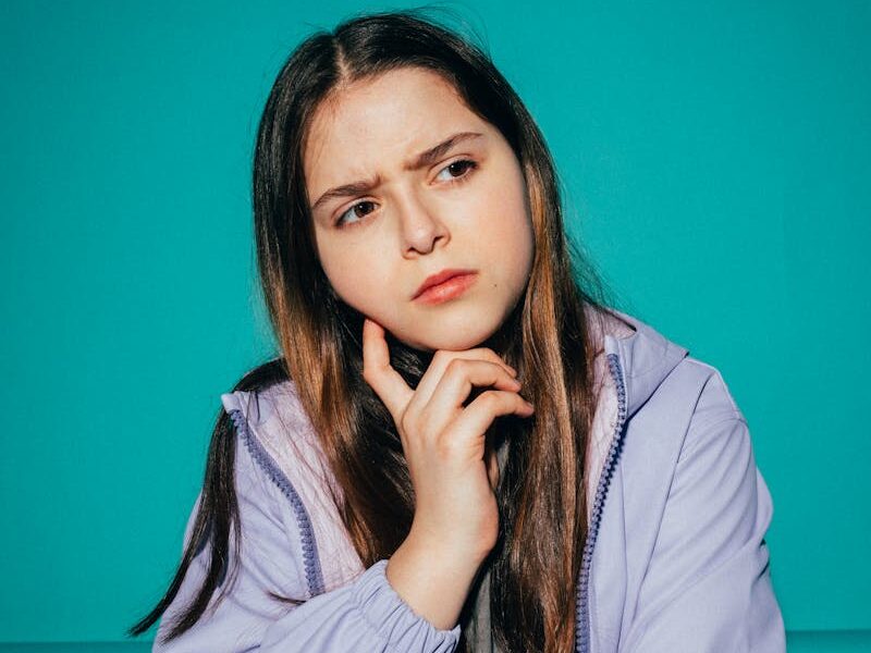 A young girl pondering deeply against a vibrant teal background.