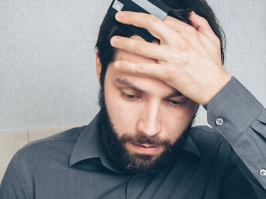 A stressed man looks at his smartphone, holding a credit card in his hand.