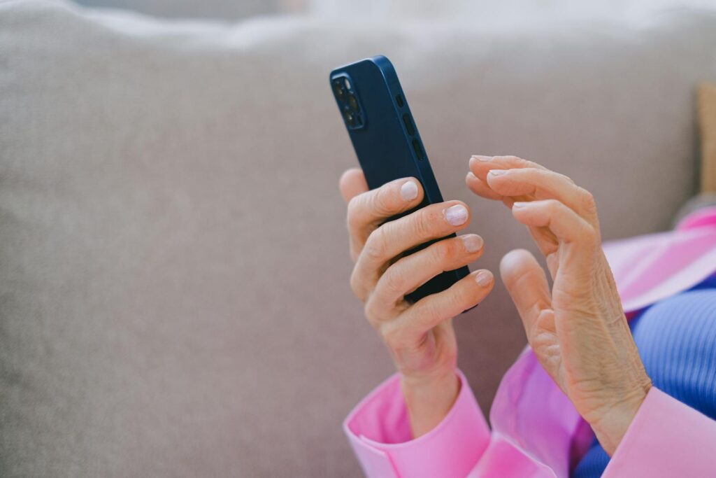 A senior adult in pink jacket using a smartphone while relaxing on a sofa indoors.