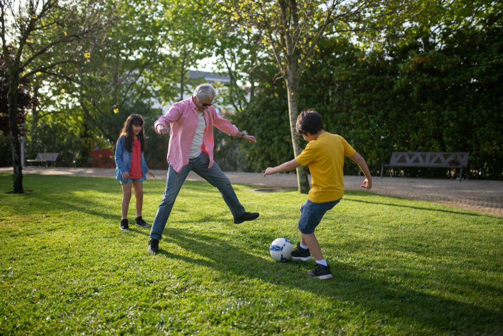 A grandfather enjoys a game of soccer with his grandchildren in a sunny park in Portugal.