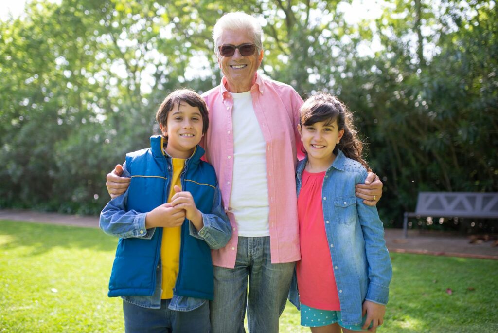 A cheerful grandfather poses with his smiling grandchildren outdoors in a sunny park.