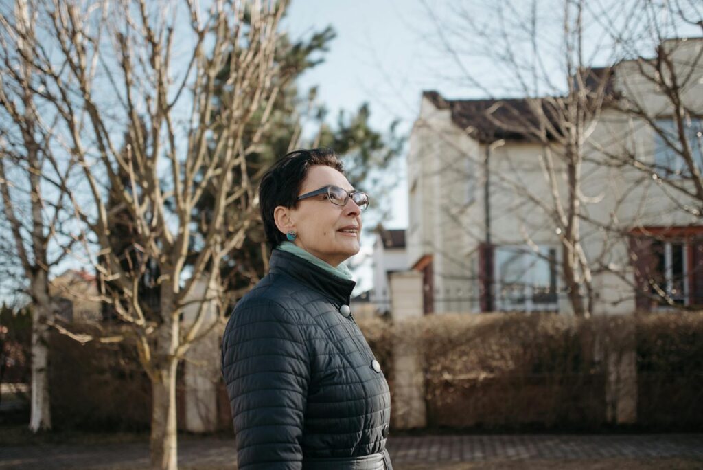 Adult woman in a winter jacket standing outdoors near urban homes, surrounded by bare trees in daylight.