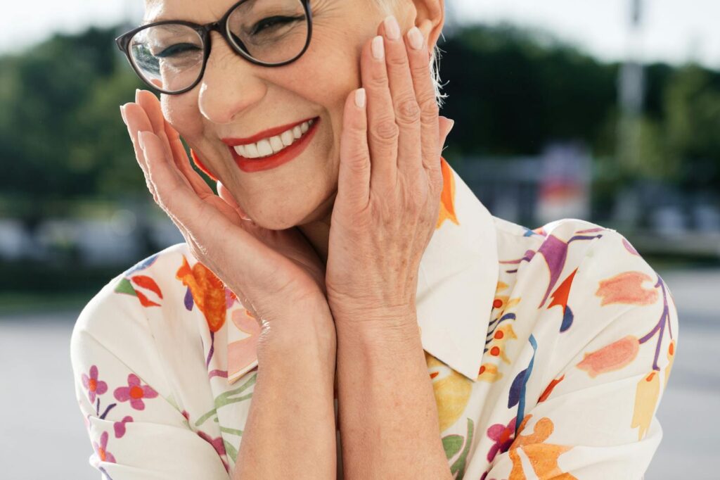 Vibrant portrait of a senior woman with glasses and a floral shirt, smiling joyfully outdoors.
