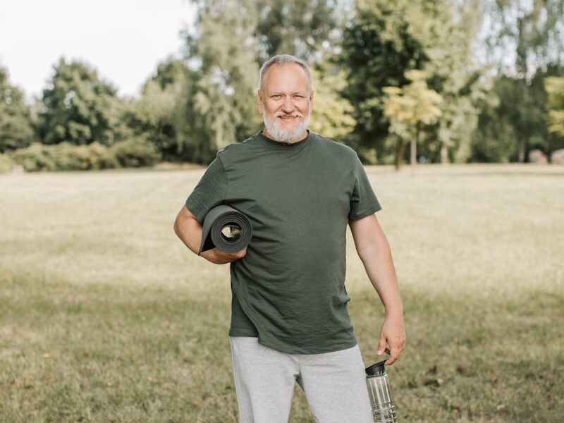 Elderly man standing outdoors, holding water bottle and yoga mat, embracing a healthy lifestyle.