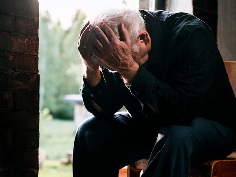 Senior man in black, grieving alone, sitting on a wooden chair by a doorway in a pensive pose.