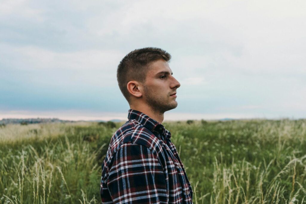 Side view portrait of a man in a plaid shirt in a green field under a cloudy sky.