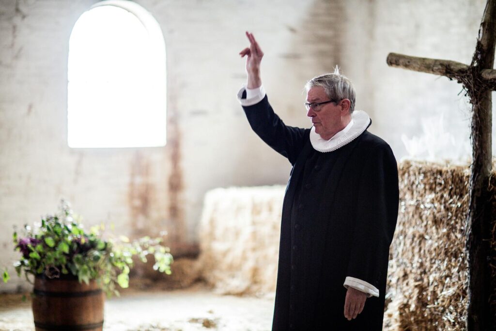 Portrait of an elderly priest conducting a ceremony in a rustic barn setting with hay bales and natural light.