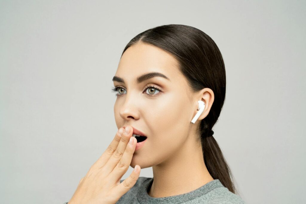 Portrait of a young woman with wireless earbuds, expressing surprise against a gray background.