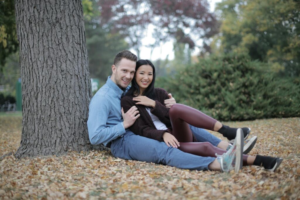 Joyful couple sitting on fallen leaves under a tree, embracing love in a serene autumn park.