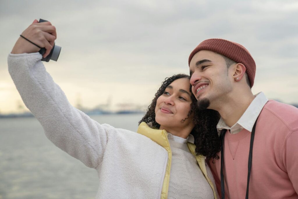 Smiling Latin American couple in warm clothes standing near river while taking self photo on analog camera in daylight under cloudy gray sky
