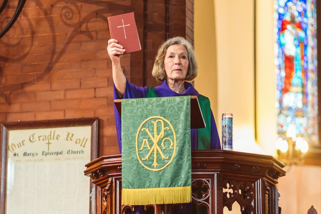 Female pastor reading from Bible during a church sermon with stained glass in background.