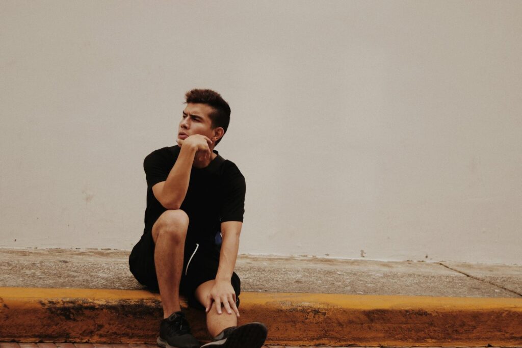 Man in casual black attire sitting thoughtfully on pavement against a white wall.